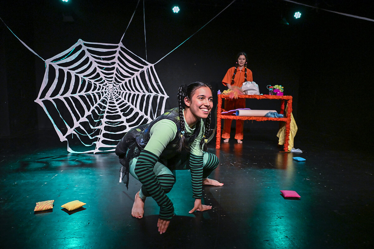 Theaterszene mit einer als Spinne kostümierten Person vor einem Spinnennetz; im Hintergrund steht eine weitere Person an einem Requisitentisch.