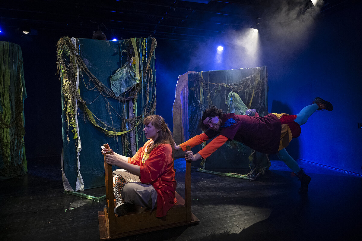 Zwei Darsteller vor einer Urwaldkulisse mit bläulichem Bühnenlicht. Die Schauspielerin sitzt auf einem umgedrehten Tisch, während der Schauspieler von hinten mit beiden Händen dranhängt.