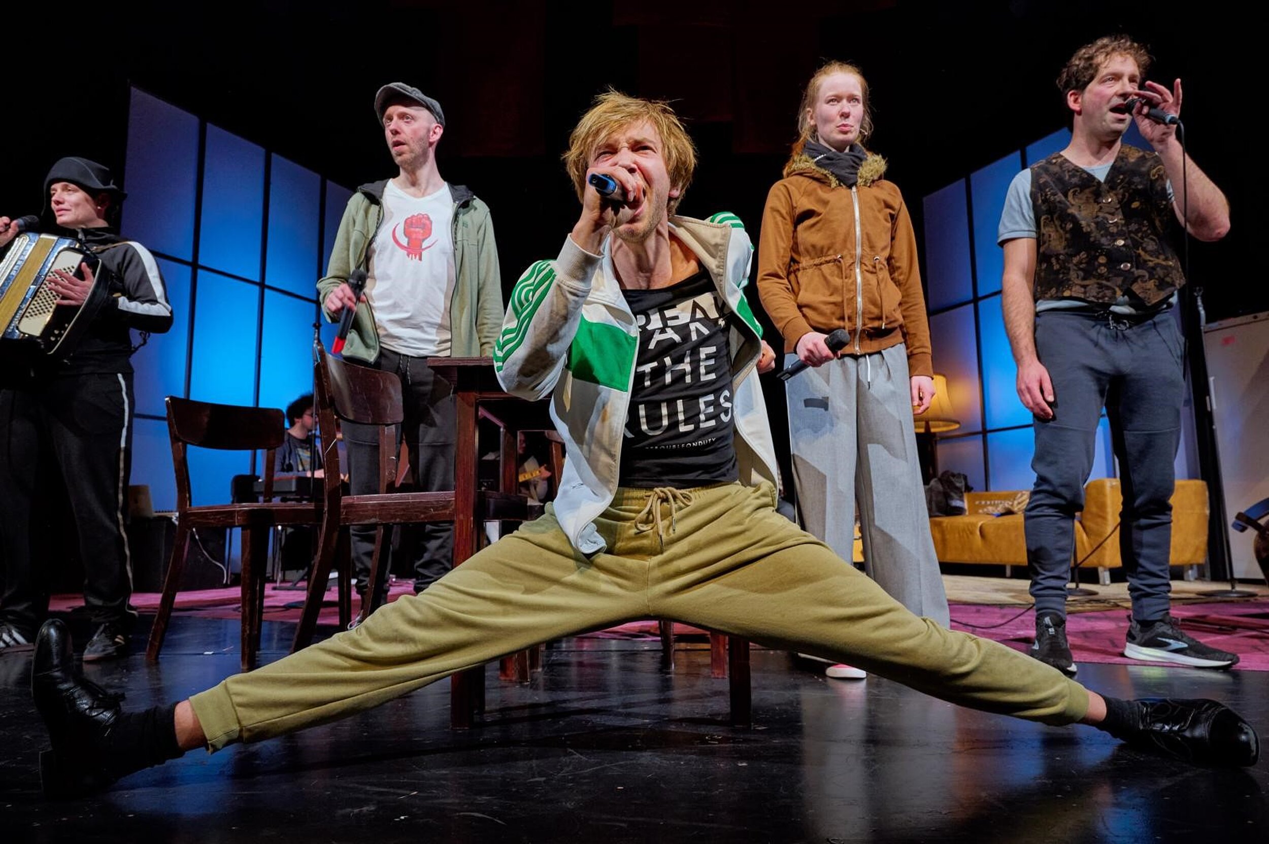 Fünf Schauspieler stehen auf einer Bühne, einer im Vordergrund macht einen Männerspagat und singt ins Mikrofon