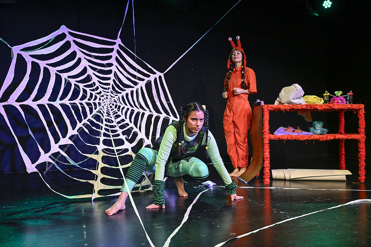 Kauernde Spinne vor einem großen Spinnennetz; im Hintergrund steht eine weitere Figur mit roter Kopfbedeckung, die auch Augen zeigen, neben einem Requisitentisch.