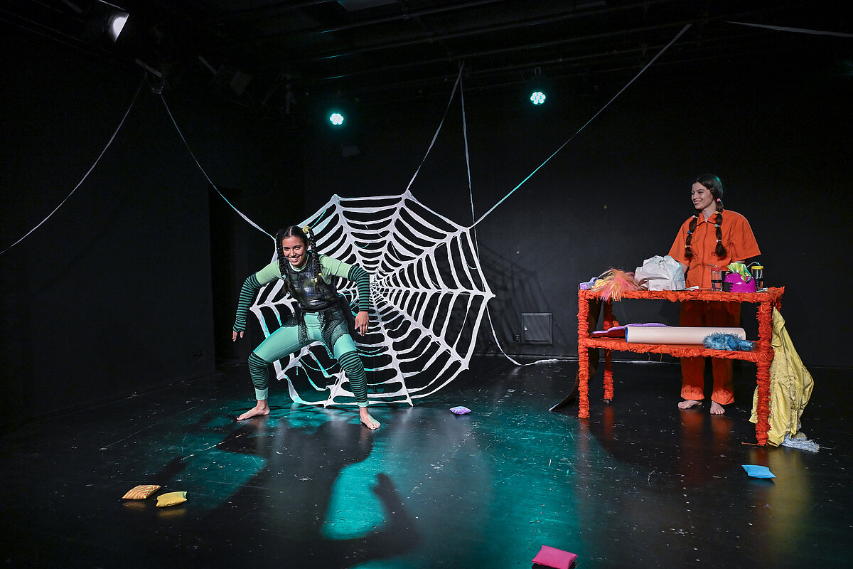 Theaterszene mit einer als Spinne kostümierten Person vor einem großen Spinnennetz; eine weitere Person steht an einem Tisch mit Requisiten auf der Bühne.