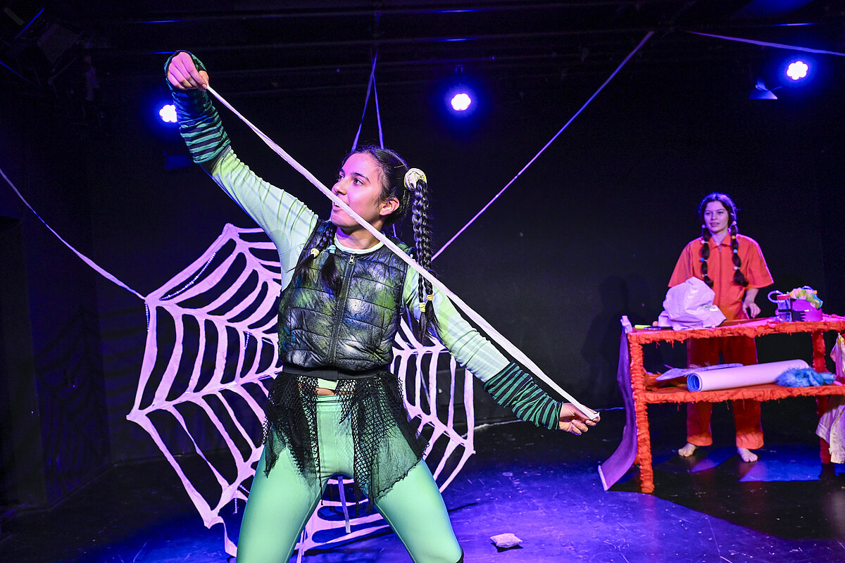 Spinne vor einem großen Spinnennetz, im Hintergrund eine weitere Figur an einem Requisitentisch.