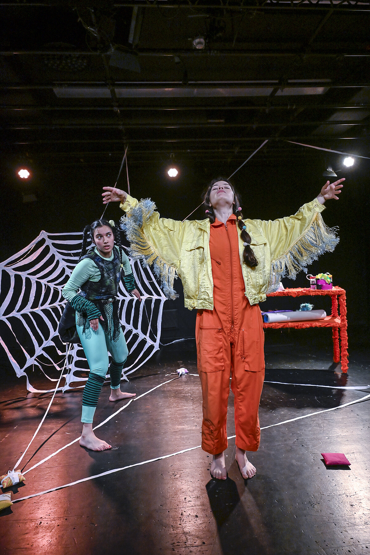 Spinne und weitere Figur, die die Hände nach oben streckt, vor einem großen Spinnennetz auf der Theaterbühne.