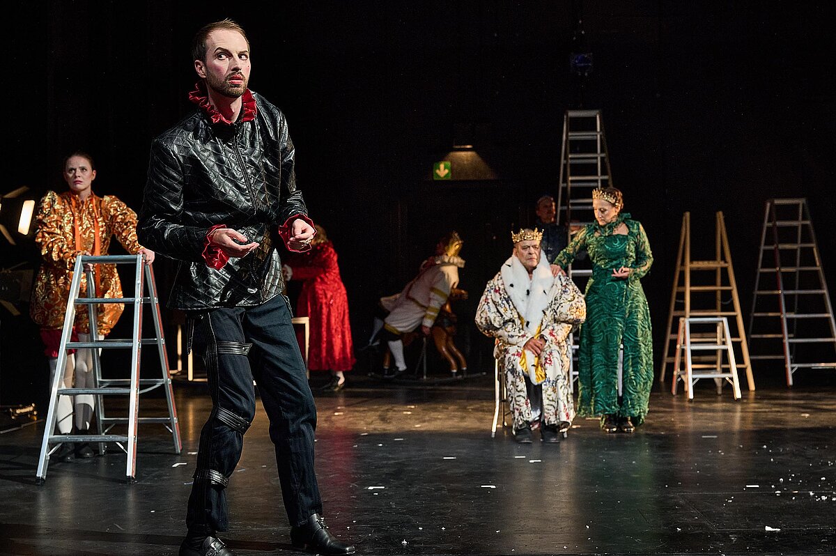 Richard III, in schwarzer Kleidung mit rotem Kragen, steht im Vordergrund der Bühne. Im Hintergrund befinden sich mehrere Darstellende in historischen Kostümen, zwei davon mit Kronen auf dem Kopf, zwischen zahlreichen Stehleitern.