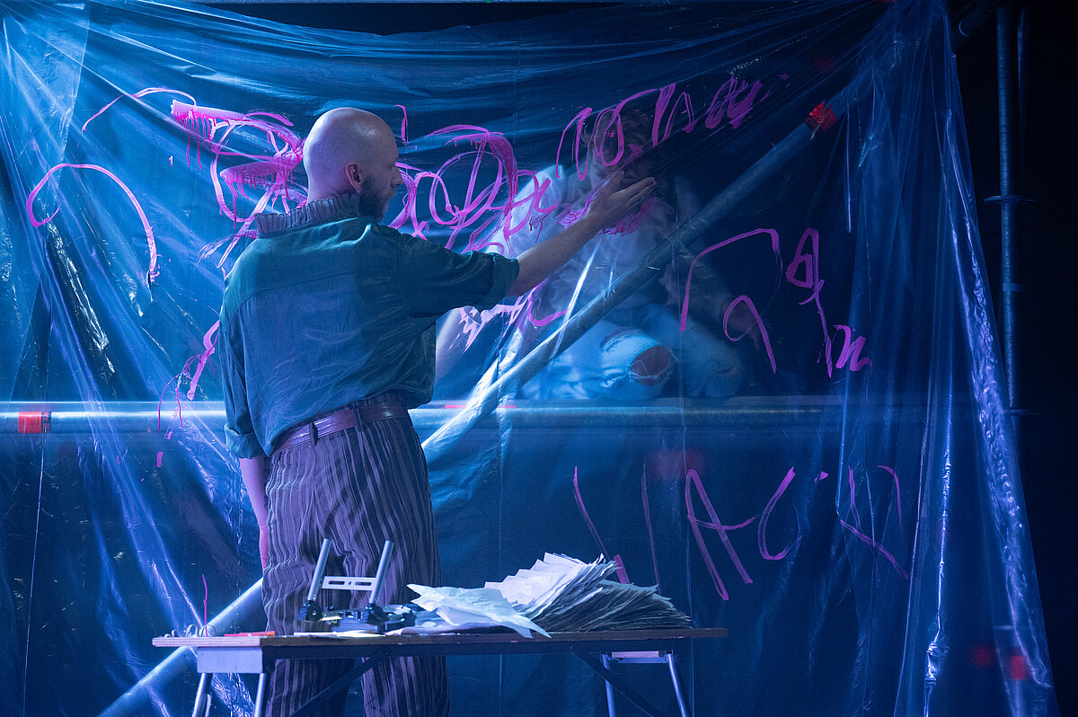 Darsteller vor transparenter, pink bekritzelter Folienwand deutet mit der Hand auf die Folie; davor steht ein Tisch mit Papierstapeln.